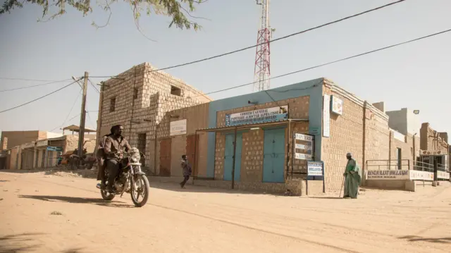 A motorcyclist rides down a dusty road in Timbuktu, with block buildings lining the street.